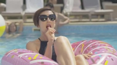 Güneş gözlüklü, su havuzunda lolipop emen kendine güvenen beyaz kadının yakın portresi. Güneşli yaz gününde, tatil köyünde donut yüzme ringinde oturan zarif, genç esmer bayan..
