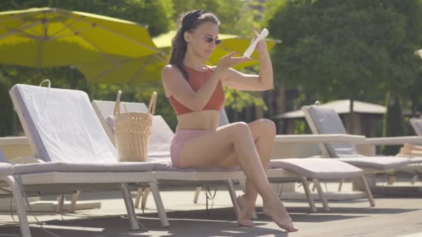 Amplia toma de hermosa mujer joven que aplica crema de bronceado de piel en las piernas y sonriendo. Retrato de chica caucásica confiada en gafas de sol que descansan en el resort en el día soleado de verano. Concepto de ocio . — Vídeo de stock