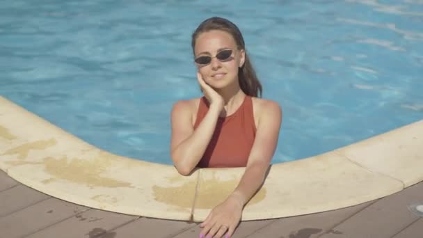 Retrato de encantadora mujer blanca alegre en gafas de sol de pie en la piscina de agua mirando a la cámara y enviando un beso. Joven hermosa chica morena descansando al aire libre en lujoso resort de verano . — Vídeos de Stock