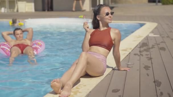 Prachtige brunette blanke vrouw met een zonnebril zuigen lolly aan het zwembad als wazig jonge man kijken naar haar van de achtergrond. Guy zit op donut zwemmen ring en bewonderen mooi meisje. — Stockvideo