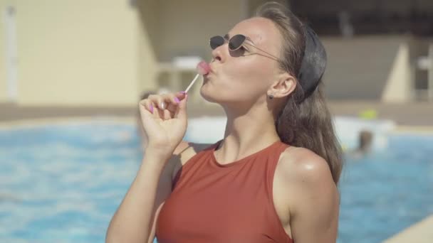 Mulher caucasiana bronzeada a chupar chupa-chupa à beira da piscina. Retrato de jovem bela menina feliz desfrutando de dia ensolarado de verão no resort de luxo. Conceito de turismo, viagens, relaxamento . — Vídeo de Stock