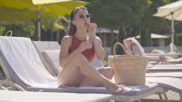 Mujer joven y relajada disfrutando de un cóctel frío sentado en el solárium del resort. Retrato de hermosa chica caucásica delgada descansando al aire libre en el soleado día de verano bebiendo bebida refrescante . — Vídeos de Stock