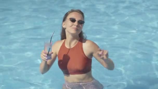 Foto en medio de una joven positiva bebiendo cóctel y bailando en el agua en la piscina. Retrato de morena hermosa chica caucásica en gafas de sol disfrutando de vacaciones en el resort en el soleado día de verano . — Vídeos de Stock