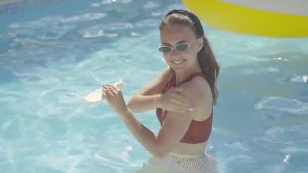 Tiro médio de menina bronzeada sorridente em óculos de sol em pé na piscina ao ar livre e aplicação de creme bronzeado no ombro. Retrato de feliz jovem turista caucasiano desfrutando de férias no resort no dia ensolarado . — Vídeo de Stock