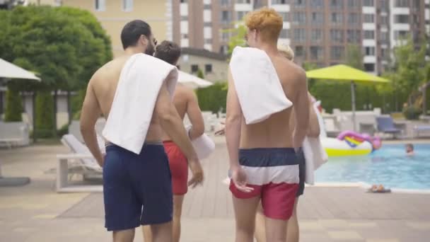 Camera suit les hommes athlétiques confiants marchant le long de la piscine dans un complexe luxueux. Vue de dos de jeunes amis masculins de différentes ethnies marchant et parlant sur la journée ensoleillée d'été en plein air. — Video