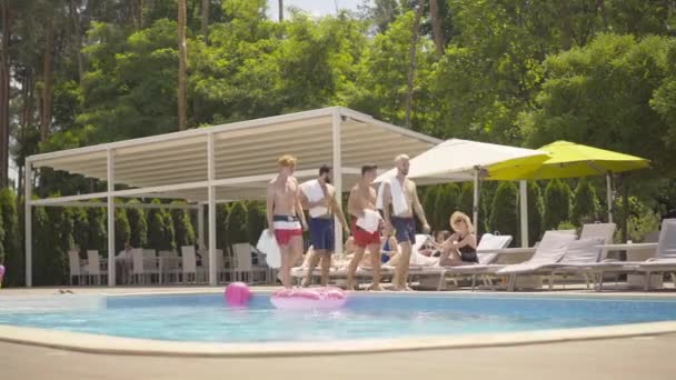 Tiro largo de quatro homens atléticos esportivos que passam por mulheres bonitas magras que se sentam em sunlounges na beira da piscina. Retrato de turistas felizes masculinos e femininos descansando no dia ensolarado no resort de luxo . — Vídeo de Stock