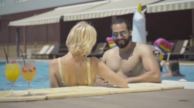Güneş gözlüklü yakışıklı Orta Doğulu genç adam su havuzunda sarışın ve beyaz bir kadınla flört ediyor. Kendine güvenen zengin turistlerin portresi yaz günü lüks tatil köylerinde dinleniyor..