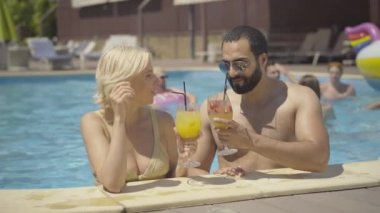 Neşeli ırklar arası çiftler bardaklarını tokuşturup bilardoda kokteyl içiyorlar. Güneşli bir günde lüks bir tatil köyünde konuşan mutlu, rahat Orta Doğulu erkek ve beyaz kadın portresi..