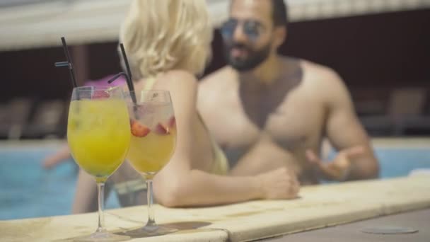 Gros plan de verres à cocktail debout au bord de la piscine avec un couple flou parlant en arrière-plan. Jeune homme confiant du Moyen-Orient et femme caucasienne au repos à la station. Relations multiraciales. — Video