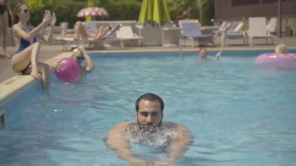 Schöner selbstbewusster Mann aus dem Nahen Osten, der zum Pool schwimmt, zwinkert und in die Kamera lächelt. Porträt junger positiver männlicher Tourist posiert an einem Sommertag im Pool eines luxuriösen Resorts. — Stockvideo