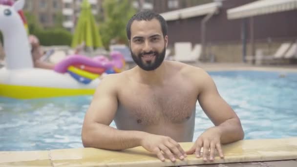 Ritratto di un bel giovane mediorientale in posa in piscina nel lussuoso resort estivo. Barbuto atletico turista maschio con sorriso dentato in piedi a bordo piscina e guardando la fotocamera. — Video Stock