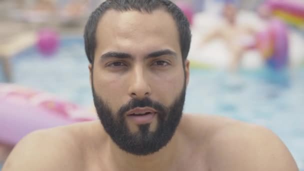 Close-up rosto de barbudo homem do Oriente Médio com olhos castanhos e cabelo preto olhando para câmera e chiclete. Retrato de confiante jovem turista posando à beira da piscina no resort ensolarado . — Vídeo de Stock