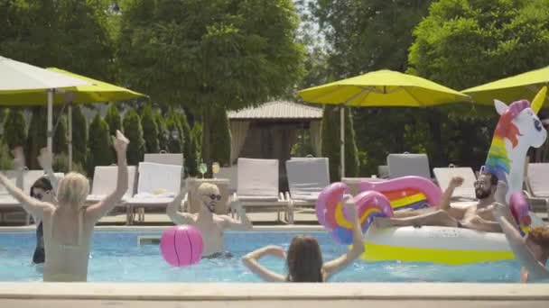 Vrolijke Kaukasische en Midden-Oosterse mannen en vrouwen spelen waterbasketbal in het zwembad. Groep gelukkige jongeren die genieten van vakantie in het resort op zonnige zomerdag buiten. — Stockvideo