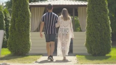 Kafkasyalı genç bir çiftin tatil köyündeki açık hava kafeye girerken geniş açılı görüntüsü. Kendinden emin esmer bir adam ve kadın güneşli bir yaz gününde yemek için restorana yürüyorlar..