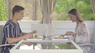 Açık hava restoranında şarküteri yiyen ve konuşan mutlu turistlerin yan görüntüsü. Genç beyaz adam ve kadın güneşli yaz gününde kafede dinleniyor ve sohbet ediyorlar. Lüks turizm.