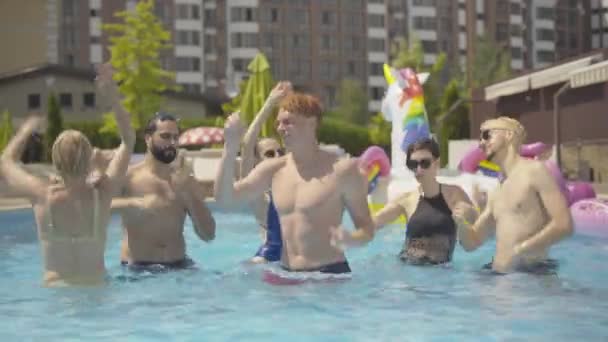 Felices turistas sonrientes bailando y salpicando agua en la piscina en un día soleado. Retrato de jóvenes alegres hombres y mujeres de Oriente Medio y el Cáucaso divirtiéndose al aire libre en un lujoso resort . — Vídeos de Stock