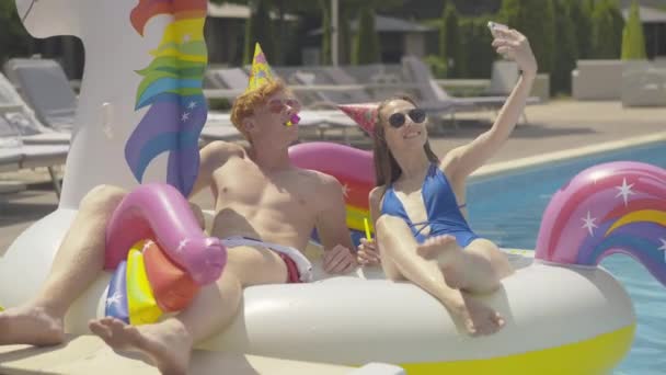 Couple caucasien riche positif prenant selfie reposant sur anneau de natation licorne dans la piscine. Portrait d'un jeune homme et d'une jeune femme souriants reposant dans un complexe luxueux par une journée ensoleillée en plein air. — Video