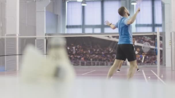 Tiro largo de dois homens a jogar badminton no campo interior. Rack foco muda para shuttlecock deitado na frente. Treinamento de desportistas atléticos em ginásio . — Vídeo de Stock