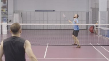 Siyah spor giyimli esmer bir adamla badminton oynayan mavi tişörtlü beyaz bir genç sporcu. Spor salonunda iç sahada antrenman yapan kendinden emin sporcuların portresi..