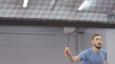 Beyaz tenli genç sporcunun portresi kapalı sahada servis yapıyor. İçeride badminton oynayan gülümseyen atletik adam. Sportif yaşam tarzı.