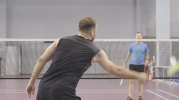 Bakåt bild av tatuerade kaukasiska idrottsman tävlar i badminton med ung suddig man i bakgrunden. Mellanslag av två män som spelar sport spel i gymmet inomhus. — Stockvideo