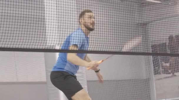 Die Live-Kamera verfolgt die Bewegung eines jungen positiven kaukasischen Sportlers, der Badminton in der Sporthalle spielt. Porträt eines bärtigen brünetten Mannes, der drinnen trainiert. Sportkonzept. — Stockvideo