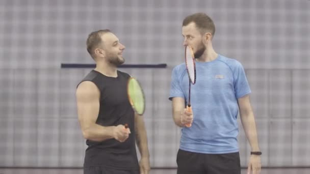 Deportistas caucásicos positivos sosteniendo raquetas de bádminton y riendo. Retrato del entrenador sonriente mostrando la posición de la mano derecha en el gimnasio. Dos hombres atléticos entrenando detrás de la red . — Vídeos de Stock