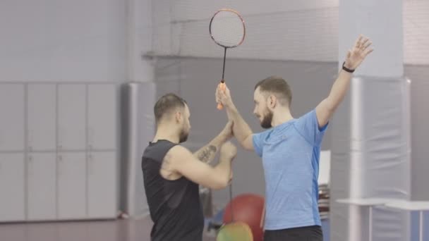 Jonge getatoeëerde coach corrigeert houding van brunette Kaukasische badmintonspeler. Portret van zelfverzekerde volwassen sporters die trainen op het binnenveld in de sportschool. — Stockvideo