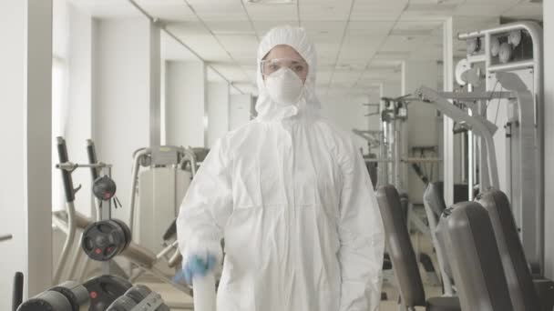 Middle shot of serious woman in protective suit and respirator spraying disinfectant in gym. Portrait of concentrated Caucasian employee posing in sports club with sanitizer. Covid-19 lifestyle. — Stock Video