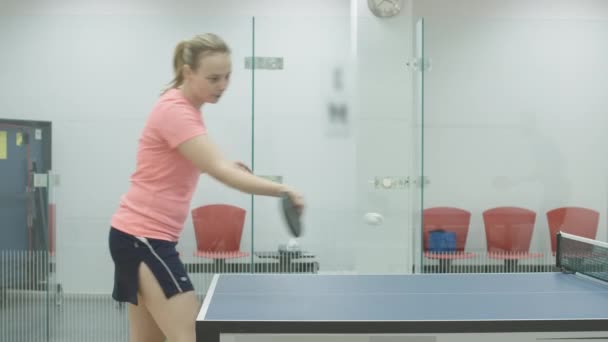 Sidovy av självsäker vit kvinna i sportkläder slå bollen med racket. Positiv ung idrottskvinna som spelar pingis i gymmet. Blond flicka träning bordtennis inomhus. — Stockvideo