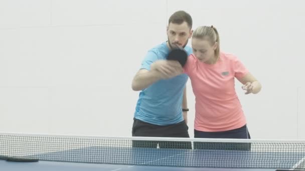 Koncentrerad kvinnlig tränare som visar höger handrörelse i pingis till ung vit man. Porträtt av professionell tränare som lär stilig skäggig idrottsman att spela bordtennis i gymmet. — Stockvideo