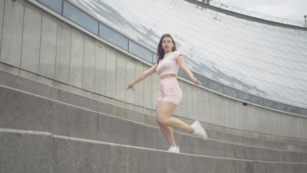 Plan large de charmante brune femme caucasienne étirant la jambe à l'extérieur. Portrait de confiant belle jeune fille à l'arrière-plan du bâtiment en verre sur les escaliers urbains. — Video