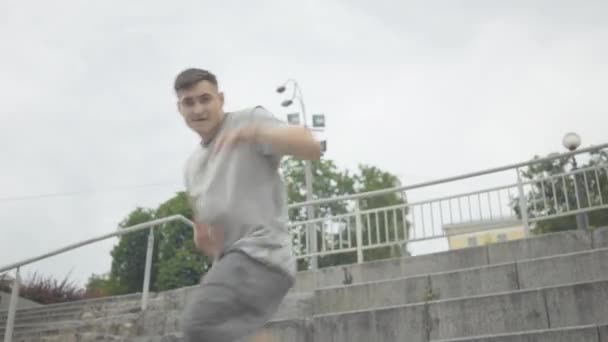 Нестримний танець упевненого молодого кавказького чоловіка на міських сходах. Портрет красивого атлетичного хлопця, який танцює з другом на вулиці в похмурий літній день. Спосіб життя, свобода. — стокове відео