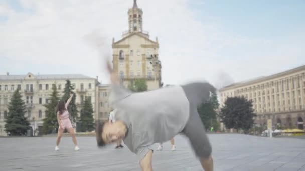 Bruna giovane uomo che esegue movimenti di danza sportiva sulla piazza della città con gli amici che ballano sullo sfondo. Ritratto di ballerino caucasico fiducioso che si gode le prestazioni sulla strada urbana. — Video Stock