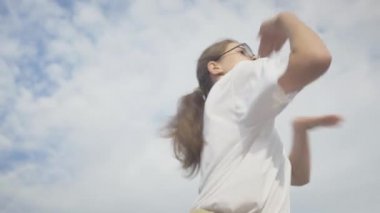 Gözlüklü beyaz bir kızın yaz gökyüzünün arka planında dans edişinin ortası. Kendine güveni tam beyaz bir kadının robot dansı figürleri..