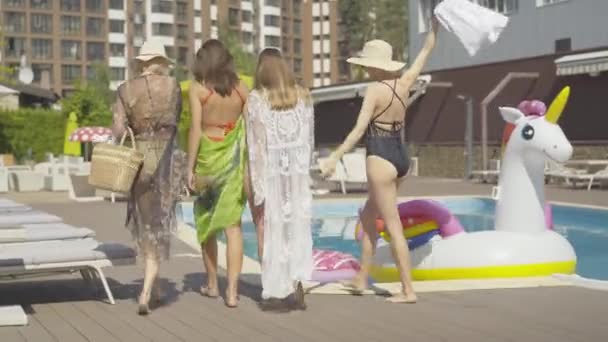 Weiträumige Rückansicht von vier kaukasischen Frauen, die am Swimmingpool des Resorts entlang laufen. Glückliche reiche Damen ruhen sich an sonnigen Sommertagen im Freien aus. Luxustourismus-Konzept. — Stockvideo