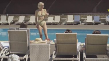Güneş gözlüklü, kendine güvenen beyaz bir kadının portresi. Güneşli salonlarda havuzun önünde yatan arkadaşlarıyla konuşuyor. Güneşli bir günde tatil köyünde dinlenen mutlu zengin turistlerin geniş bir görüntüsü.
