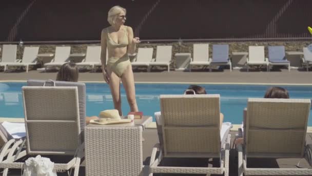 Güneş gözlüklü, kendine güvenen beyaz bir kadının portresi. Güneşli salonlarda havuzun önünde yatan arkadaşlarıyla konuşuyor. Güneşli bir günde tatil köyünde dinlenen mutlu zengin turistlerin geniş bir görüntüsü. — Stok video