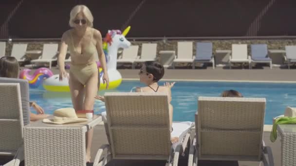 Mujer alegre en traje de baño dorado y gafas de sol poniéndose sombrero de sol y hablando con amigos tumbados en tumbonas. Retrato de turistas mujeres positivas descansando en el balneario de verano en un día soleado . — Vídeos de Stock