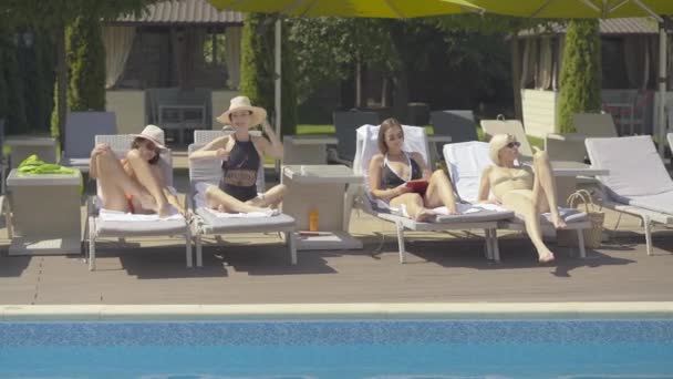 Quatro mulheres caucasianas felizes confiantes descansando em sunlounges no resort de verão. Descanso luxuoso de mulheres adultas positivas no dia ensolarado. Ampla foto de turistas relaxados à beira da piscina . — Vídeo de Stock