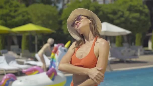 Средний снимок красивой белой женщины, использующей солнцезащитный крем на курорте. Портрет счастливого уверенного туриста, растирающего крем для загара у бассейна в солнечный летний день. Богатая женщина наслаждается отпуском. — стоковое видео