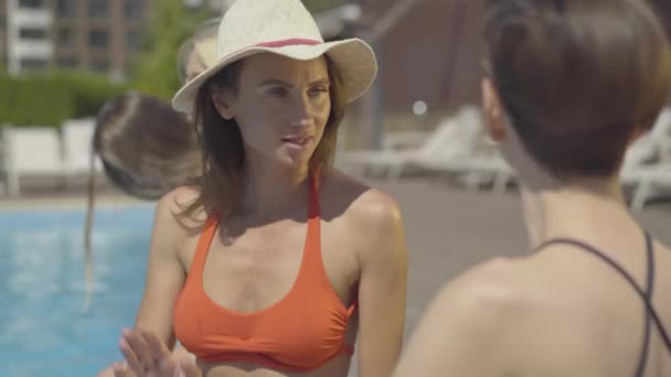 Retrato de la hermosa mujer adulta caucásica en sombrero de sol hablando con un amigo sentado junto a la piscina. Turistas ricos confiados chismorreando en el resort en un día soleado . — Vídeos de Stock