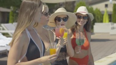 Güneş gözlüklü ve güneşli yaz gününde kokteyl içen mutlu beyaz kadınların yan görüntüsü. Kendine güvenen yetişkin turistler lüks tatil beldelerinde tatilin tadını çıkarıyorlar..