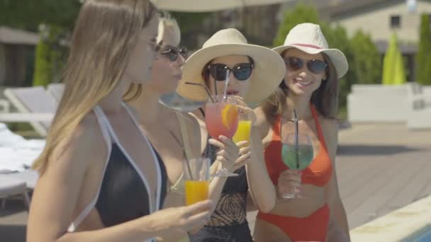Vista lateral de las mujeres caucásicas felices en gafas de sol tintineo vasos y cócteles de beber en el día soleado de verano. Turistas adultos seguros disfrutando de vacaciones en un resort de lujo . — Vídeo de stock