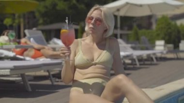 Havuz başındaki lüks bir otelde kokteyl içen kendine güvenen, gevşemiş sarışın kadının portresi. Güneş gözlüklü güzel beyaz turistin manzarası güneşli bir günde alkolün tadını çıkarıyor.