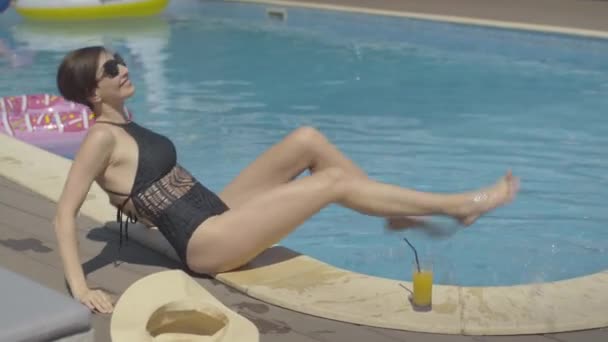 Вид сбоку на веселую брюнетку, развлекающуюся, сидя у бассейна. Портрет красивой белой женщины, плескающейся водой и улыбающейся. Счастливый турист отдыхает в солнечный летний день на курорте. — стоковое видео
