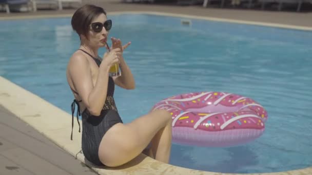 Mulher caucasiana sensual magro em óculos de sol bebendo coquetel e salpicando água na piscina no dia ensolarado. Retrato de morena bela turista descansando no resort de luxo . — Vídeo de Stock