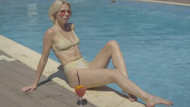 Selbstbewusst braun gebrannte blonde Frau in goldenem Badeanzug sonnt sich im luxuriösen Resort. Breitbild-Porträt eines wunderschönen erwachsenen kaukasischen Touristen mit Sonnenbrille, der an einem sonnigen Sommertag am Pool sitzt. — Stockvideo