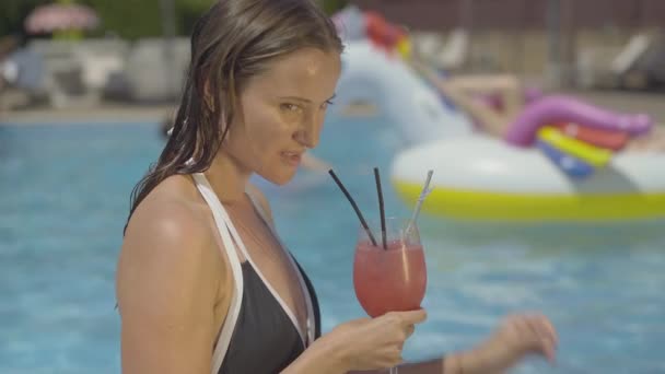 Charmante brune femme blanche aux cheveux mouillés dégustant une boisson alcoolisée au bord de la piscine le jour d'été ensoleillé. Portrait latéral d'une touriste souriante heureuse se reposant au resort. — Video