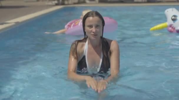 Brunette Femme caucasienne aux yeux marron nageant à la caméra dans la piscine le jour ensoleillé. Portrait de touriste adulte confiante posant au bord de la piscine dans une luxueuse station estivale. — Video